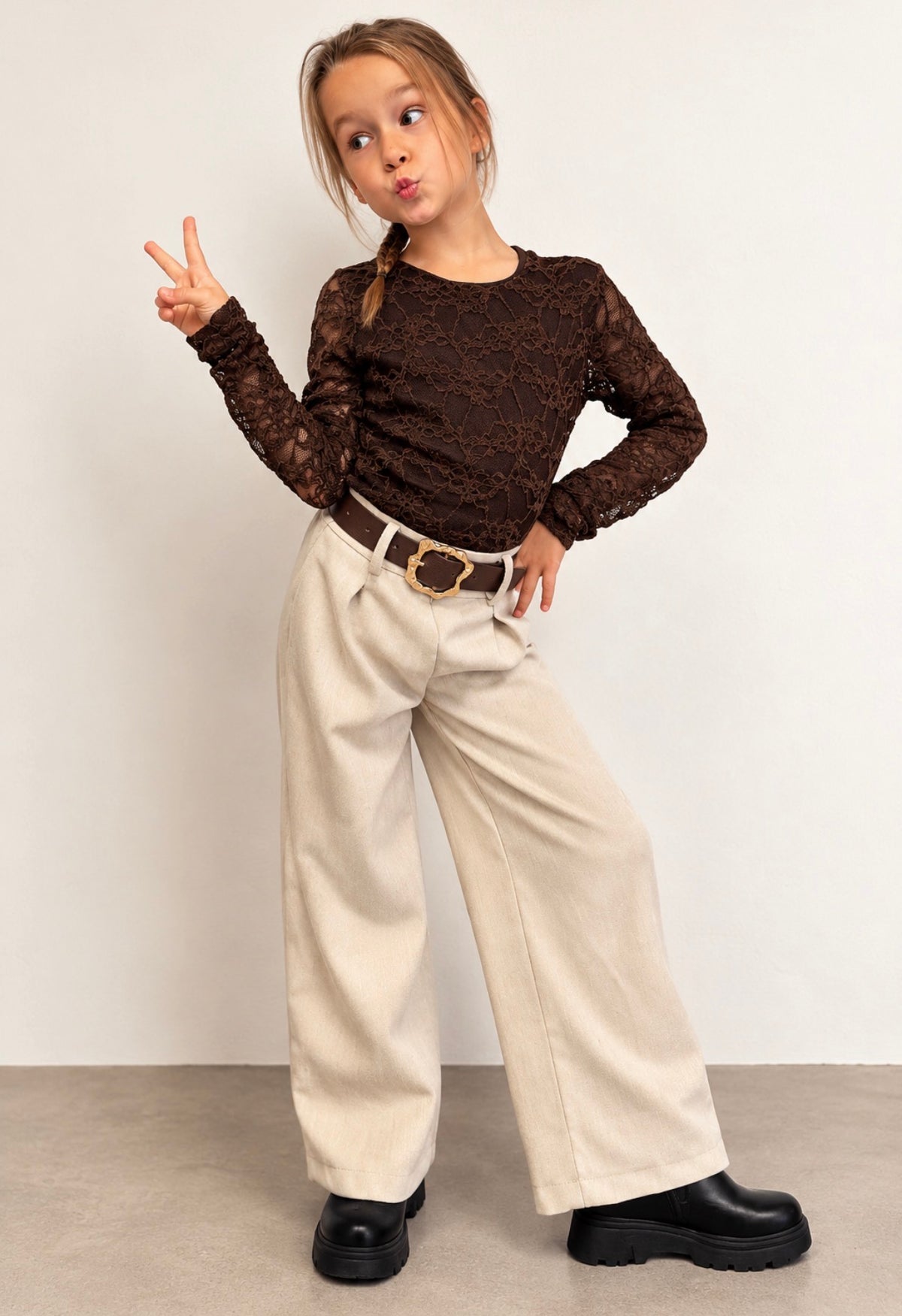 Meisje draagt een bruine kanten top met bloemenpatroon en lange mouwen, gecombineerd met een beige wide-leg pantalon.